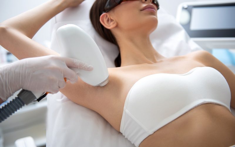 Aesthetic body treatment. Close up portrait of young relaxed beautiful woman in white underwear lying on couch and having laser hair removal procedure of armpit